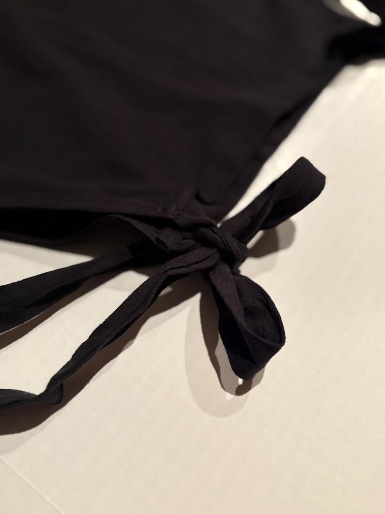 Op de afbeelding: Close-up van een zwart kledingstuk met een strikdetail. De stof lijkt een zacht, vloeiend materiaal te zijn. De strik is geknoopt en de uiteinden van de banden zijn zichtbaar. De achtergrond is licht en neutraal.