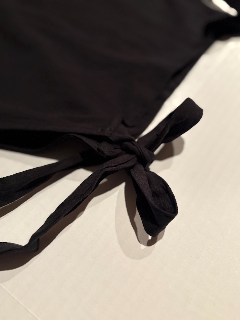 May include: A black garment featuring a fabric tie detail at the corner. The tie is knotted into a bow, with two long, loose fabric strips extending downward from the knot. The fabric appears smooth and solid black in color throughout the piece.