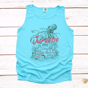 May include: A light blue tank top featuring a graphic design with a T-Rex, a red and white Jurassic Park sign, and a red SUV. The word "Jurassic" is written in red script.