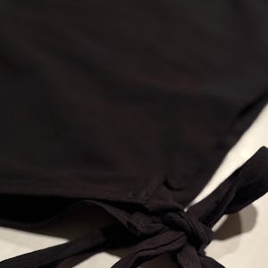 Op de afbeelding: Close-up van een zwart kledingstuk met een geknoopt detail. De stof lijkt zacht en vloeiend te zijn, met een subtiele glans. De knoop is gecentreerd aan de rechterkant van de afbeelding, met de stof die naar beneden valt.