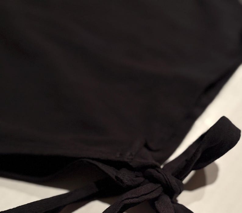 Op de afbeelding: Close-up van een zwart kledingstuk met een geknoopt detail. De stof lijkt zacht en vloeiend te zijn, met een subtiele glans. De knoop is gecentreerd aan de rechterkant van de afbeelding, met de stof die naar beneden valt.