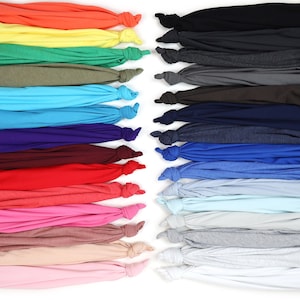 K&ouml;nnte beinhalten: Eine Auswahl an farbenfrohen T-Shirts, die am unteren Ende geknotet sind. Die Shirts sind in Reihen angeordnet und zeigen eine Vielzahl von Farbt&ouml;nen, darunter Orange, Gelb, Gr&uuml;n, Blau, Lila, Rot, Rosa und neutrale T&ouml;ne. Der Hintergrund ist wei&szlig;.