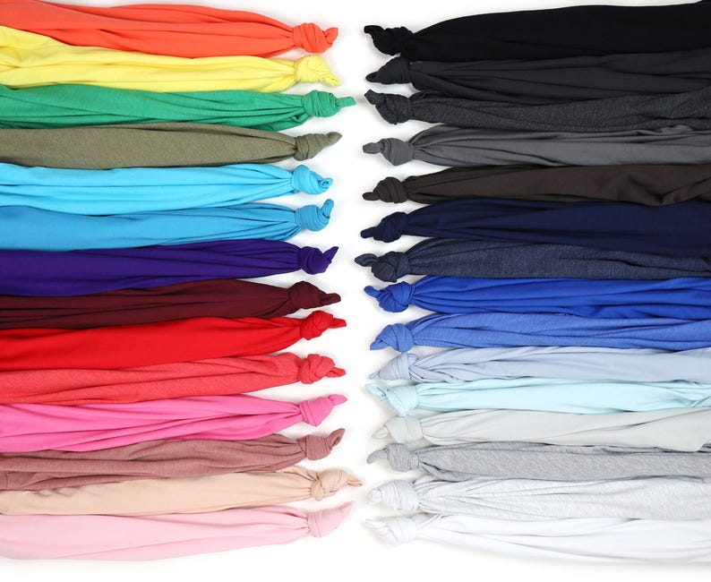K&ouml;nnte beinhalten: Eine Auswahl an farbenfrohen T-Shirts, die am unteren Ende geknotet sind. Die Shirts sind in Reihen angeordnet und zeigen eine Vielzahl von Farbt&ouml;nen, darunter Orange, Gelb, Gr&uuml;n, Blau, Lila, Rot, Rosa und neutrale T&ouml;ne. Der Hintergrund ist wei&szlig;.