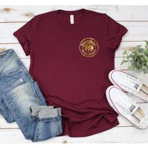 K&ouml;nnte beinhalten: Ein weinrotes T-Shirt mit einem goldenen Disneyland Railroad Logo, kombiniert mit blauen Jeans und wei&szlig;en Turnschuhen. Das Shirt steht im Mittelpunkt, mit dem Logo deutlich sichtbar. Das l&auml;ssige Outfit ist auf einer hellen Holzmaserung angeordnet.