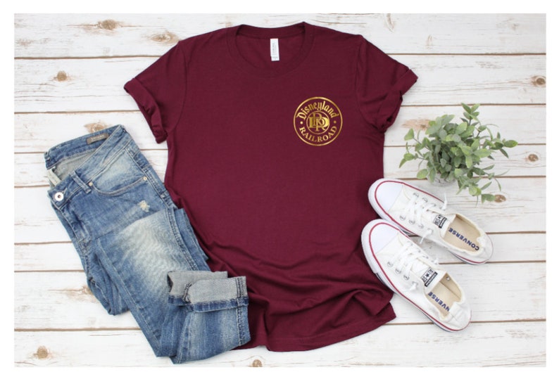 K&ouml;nnte beinhalten: Ein weinrotes T-Shirt mit einem goldenen Disneyland Railroad Logo, kombiniert mit blauen Jeans und wei&szlig;en Turnschuhen. Das Shirt steht im Mittelpunkt, mit dem Logo deutlich sichtbar. Das l&auml;ssige Outfit ist auf einer hellen Holzmaserung angeordnet.