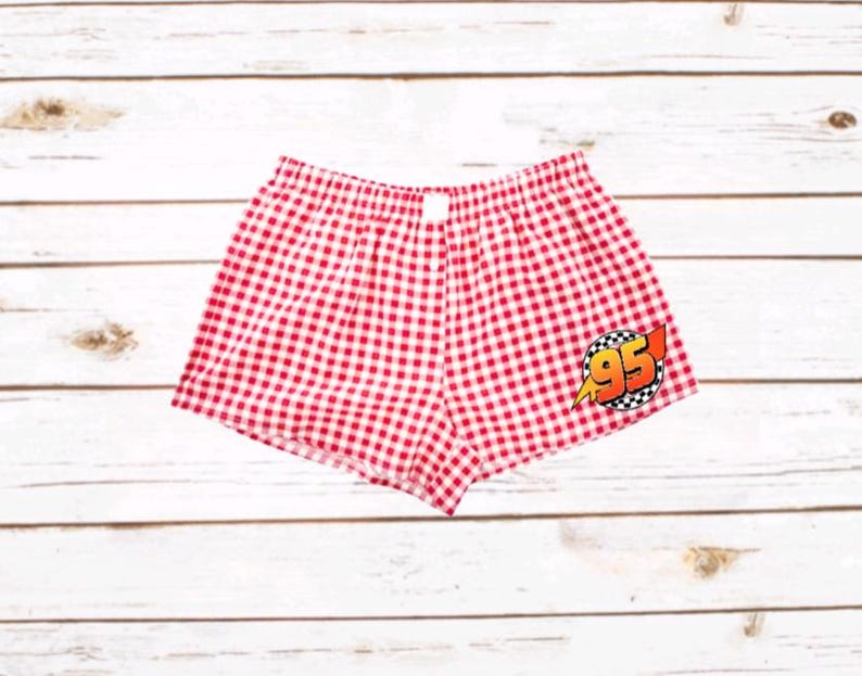 K&ouml;nnte beinhalten: Rot-wei&szlig; karierte Boxershorts mit elastischem Bund und einem grafischen Aufn&auml;her auf der unteren rechten Seite. Der Aufn&auml;her zeigt die Zahl 95 in Orange mit einem gelben Blitz und einem schwarz-wei&szlig; karierten Randdesign.