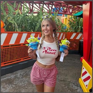 K&ouml;nnte beinhalten: Ein wei&szlig;es &auml;rmelloses bauchfreies Oberteil mit rotem Pizza Planet-Logo, rot-wei&szlig; karierte Shorts und ein gr&uuml;ner Alien-Haarreif. In jeder Hand wird ein kleines gr&uuml;nes, drei&auml;ugiges Alien-Pl&uuml;schtier mit blauem Unterteil und wei&szlig;em Etikett gehalten.