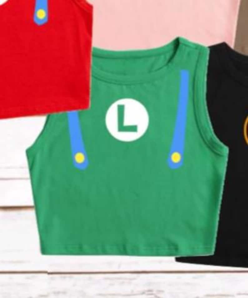 May include: Green crop top with a white "L" emblem, blue suspenders, and yellow accents. The sleeveless shirt is part of a costume set. Other shirts in the image are red, pink, and black.