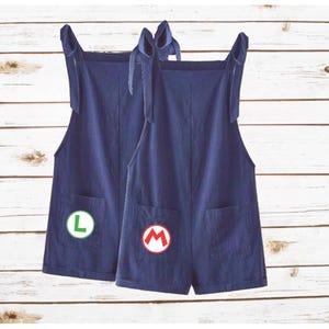 May include: Two dark blue short overalls with tie-up shoulder straps and front pockets. One pair features a circular patch with a green letter L, and the other pair features a circular patch with a red letter M on the front pocket area.