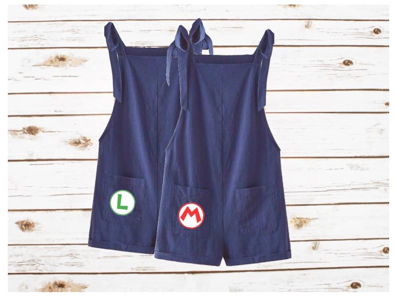 May include: Two dark blue short overalls with tie-up shoulder straps and front pockets. One pair features a circular patch with a green letter L, and the other pair features a circular patch with a red letter M on the front pocket area.