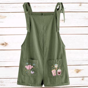 Op de afbeelding: Olijfgroene romper met geborduurde zakdetails. De romper heeft verstelbare strikbandjes en twee voorzakken met kleurrijke geborduurde ontwerpen van een ballon, popcorn, ijs en een drankje.