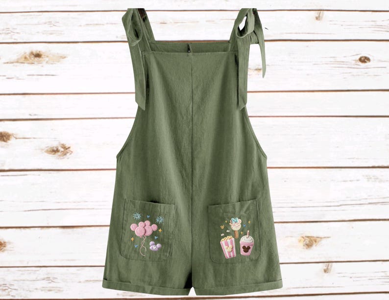 Op de afbeelding: Olijfgroene romper met geborduurde zakdetails. De romper heeft verstelbare strikbandjes en twee voorzakken met kleurrijke geborduurde ontwerpen van een ballon, popcorn, ijs en een drankje.