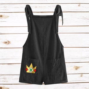 May include: Black short overalls featuring tie-shoulder straps and two front pockets. The left pocket displays an embroidered yellow crown graphic with a green center gem and red side accents, providing a distinct contrast against the black fabric.