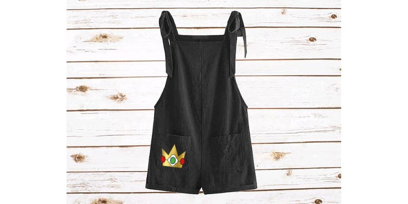 May include: Black short overalls featuring tie-shoulder straps and two front pockets. The left pocket displays an embroidered yellow crown graphic with a green center gem and red side accents, providing a distinct contrast against the black fabric.