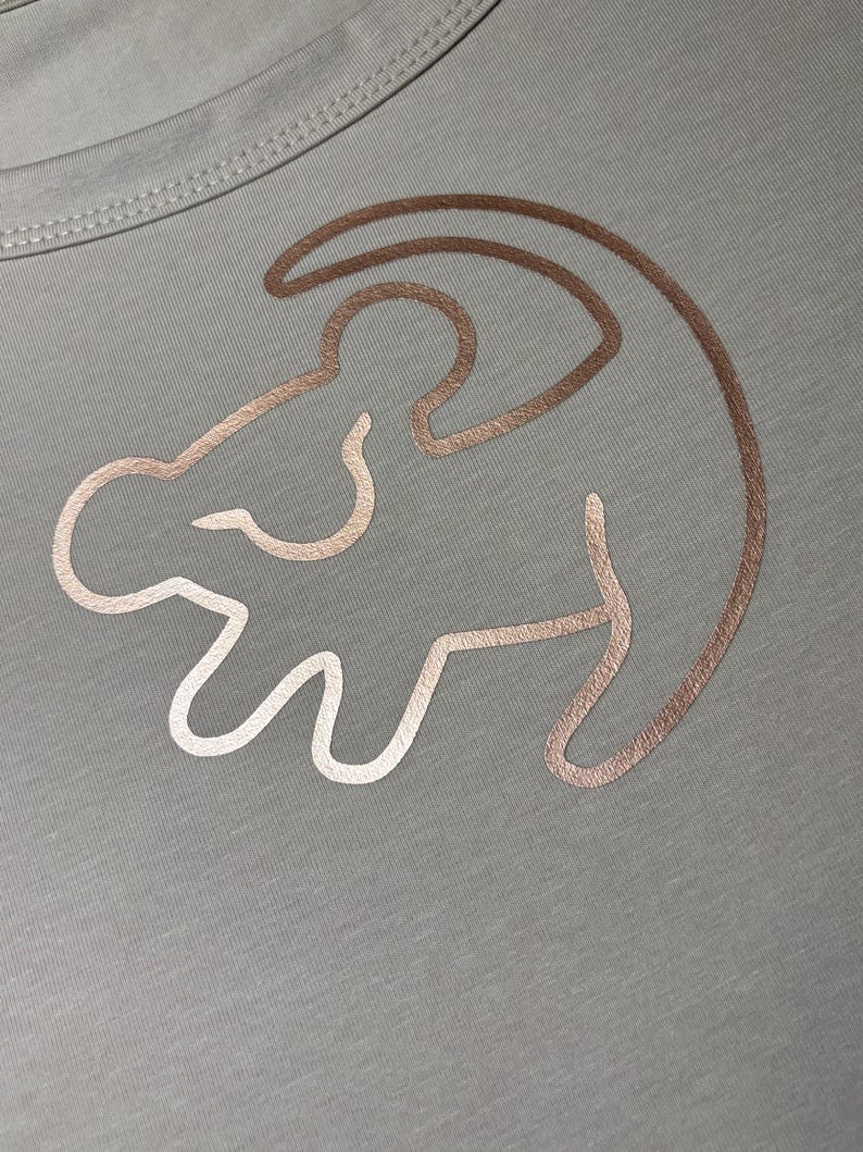 May include: A light gray t-shirt with a gold outline of Simba from The Lion King.