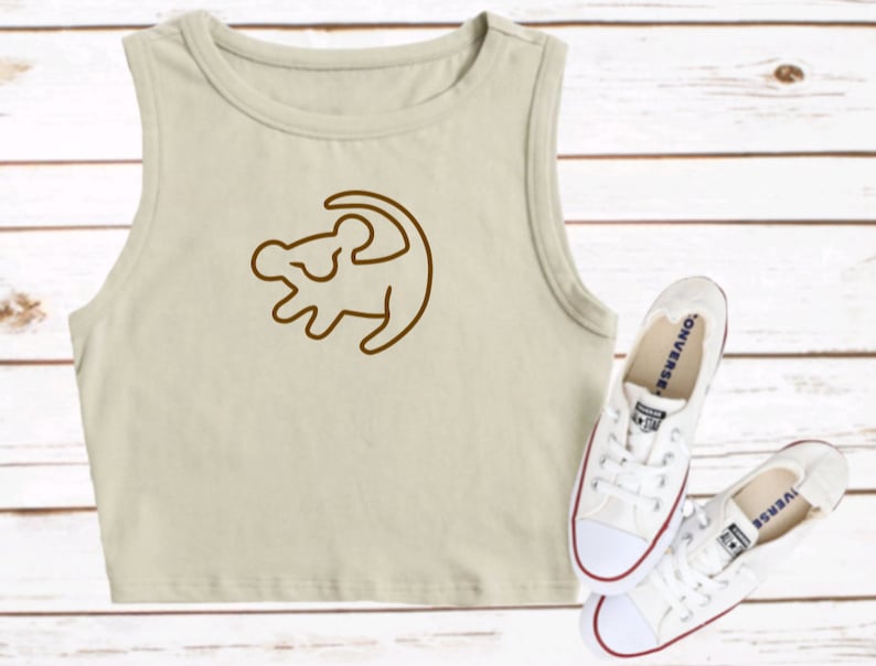 May include: A beige sleeveless crop top with a brown outline of Simba's profile from The Lion King. The top is paired with white sneakers.