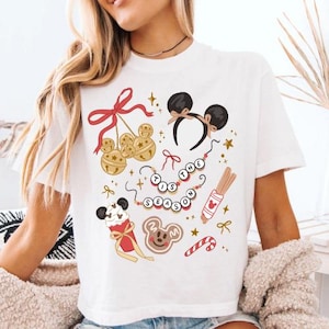 May include: White t-shirt featuring a festive design with Mickey Mouse ears, bells, and gingerbread cookies. The phrase "'Tis the Season" is displayed. The design includes red bows, stars, and candy canes, perfect for the holiday season.