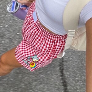 K&ouml;nnte beinhalten: Rot-wei&szlig; karierte Shorts mit elastischem Bund, kombiniert mit einem schlichten wei&szlig;en Kurzarm-Oberteil, einem beigen Rucksack und einer hellvioletten Isolierflasche. Eine kleine Grafik einer Schreibtischlampe und eines Balls ist zu sehen.
