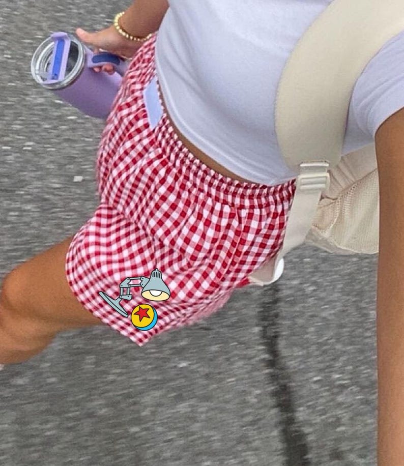 K&ouml;nnte beinhalten: Rot-wei&szlig; karierte Shorts mit elastischem Bund, kombiniert mit einem schlichten wei&szlig;en Kurzarm-Oberteil, einem beigen Rucksack und einer hellvioletten Isolierflasche. Eine kleine Grafik einer Schreibtischlampe und eines Balls ist zu sehen.