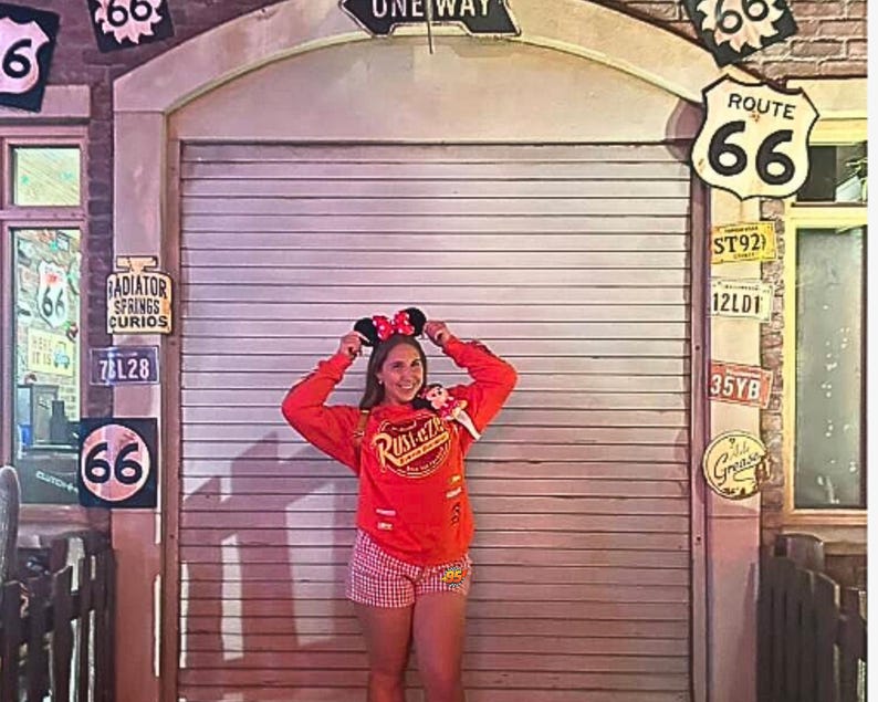 K&ouml;nnte beinhalten: Ein leuchtend orangefarbenes Langarm-Sweatshirt mit dem gelb-roten Rust-eze-Logo, kombiniert mit rot-wei&szlig; karierten Shorts. Das Outfit wird durch schwarze Mausohren mit einer rot-wei&szlig; gepunkteten Schleife erg&auml;nzt.