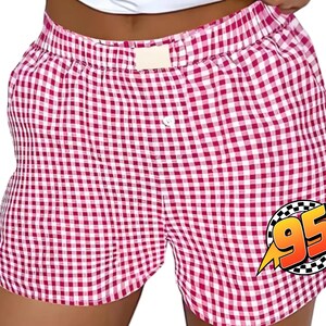K&ouml;nnte beinhalten: Boxershorts mit rot-wei&szlig;em Vichy-Karo-Muster, elastischem Bund und einem einzelnen wei&szlig;en Knopf in der Mitte. Eine kreisf&ouml;rmige Grafik mit kariertem Rand zeigt die orangefarbene Zahl 95 mit einem gelben Blitz auf der rechten Seite.