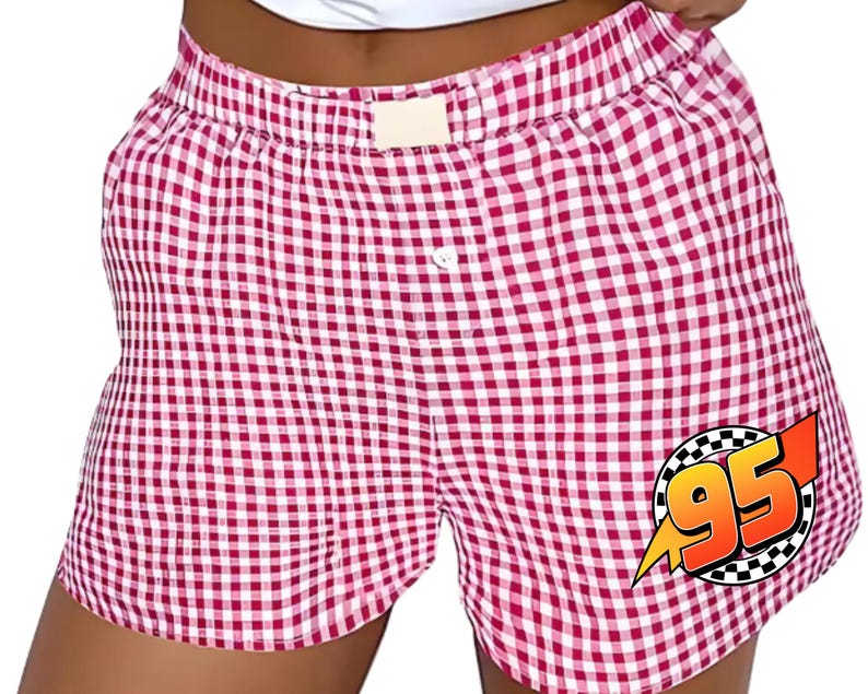 K&ouml;nnte beinhalten: Boxershorts mit rot-wei&szlig;em Vichy-Karo-Muster, elastischem Bund und einem einzelnen wei&szlig;en Knopf in der Mitte. Eine kreisf&ouml;rmige Grafik mit kariertem Rand zeigt die orangefarbene Zahl 95 mit einem gelben Blitz auf der rechten Seite.