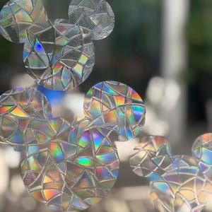 May include: Iridescent window clings in the shape of Mickey Mouse heads. The clings are made of a material that reflects a rainbow of colors. The shapes are arranged in a cluster, creating a playful and decorative effect.