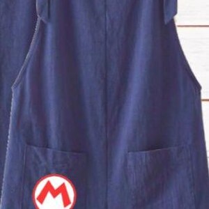 May include: A pair of dark blue denim overalls featuring two front pockets and shoulder straps. The left pocket displays a circular white patch with a red letter M outlined in red, resembling a classic video game character emblem.