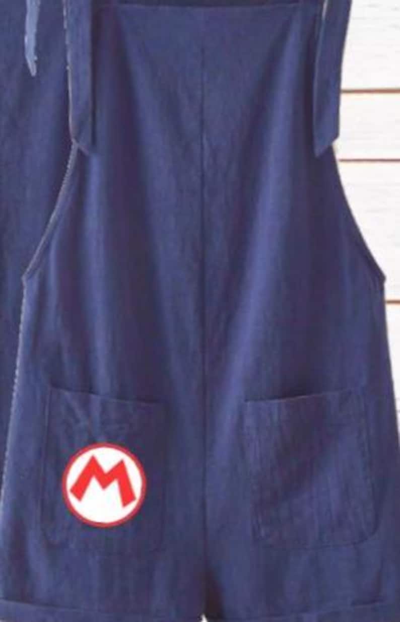 May include: A pair of dark blue denim overalls featuring two front pockets and shoulder straps. The left pocket displays a circular white patch with a red letter M outlined in red, resembling a classic video game character emblem.