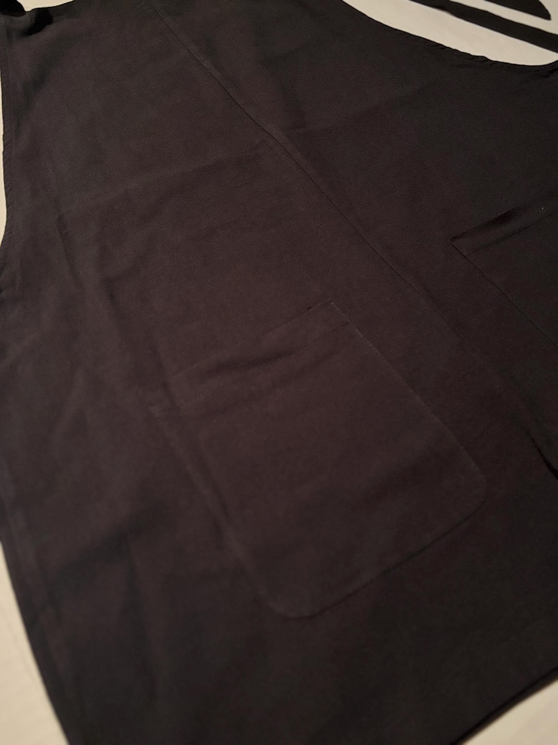 Op de afbeelding: Een donkergrijze schort met twee grote voorzakken. De schort heeft een eenvoudig, functioneel ontwerp, met een slab en bandjes. De stof lijkt een stevig, geweven materiaal te zijn, geschikt voor keuken of werkplaats.