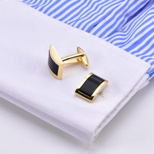 Puede incluir: Un par de gemelos dorados con una incrustación rectangular negra. Se muestran sobre un puño de camisa blanca con tela a rayas azules y blancas en el fondo. Los gemelos tienen un diseño clásico y elegante.
