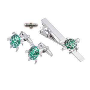 May include: A set of silver-toned cufflinks and tie bar featuring green enamel turtle designs. The tie bar has the words "I love you" engraved on it.