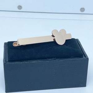 May include: Gold-toned tie bar with a Mickey Mouse head design. The tie bar is displayed on a black velvet box.