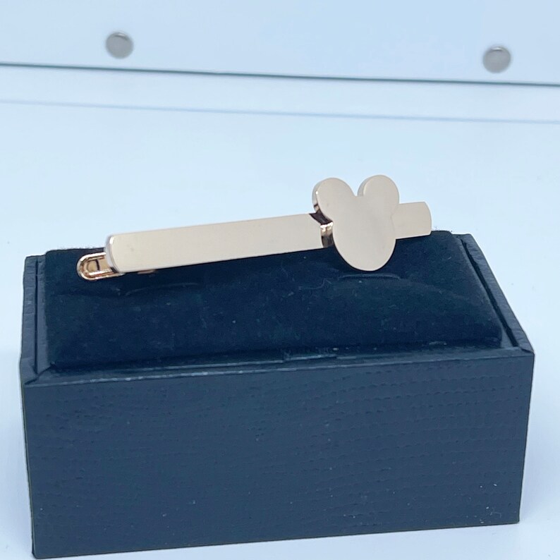 May include: Gold-toned tie bar with a Mickey Mouse head design. The tie bar is displayed on a black velvet box.