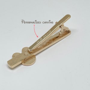 May include: Gold-toned tie clip with a clover design. The clip has a personalized inscription that reads "A&R 24.5.18".