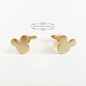 May include: A pair of gold-coloured cufflinks shaped like a mouse head. The cufflinks are set against a white background. The text "personalized signature" is above the cufflinks.