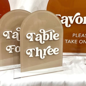 May include: Two acrylic table number signs with rounded tops. The signs are beige with white text. The text on the left sign reads "Table Four". The text on the right sign reads "Table Three".
