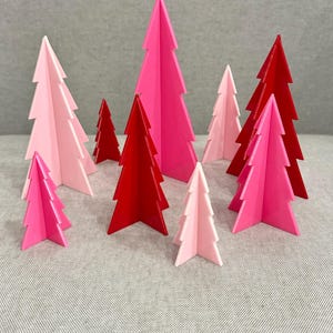 以下が含まれることがあります： ピンクと赤の様々な色合いの、装飾的で幾何学的なクリスマスツリーのコレクション。木は灰色の表面に配置され、お祝いのディスプレイを作成します。木はしっかりとした素材でできており、シンプルでモダンなデザインです。