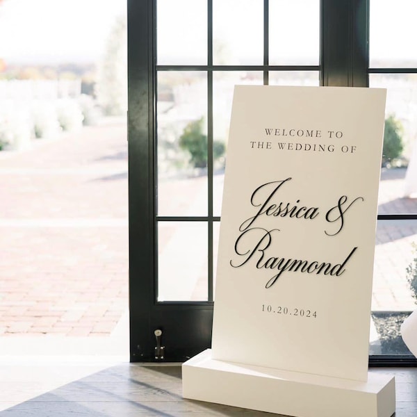 Puede incluir: Un letrero de bienvenida de boda rectangular de color blanco sobre una base blanca a juego. El letrero presenta texto en negro que dice WELCOME TO THE WEDDING OF, seguido de los nombres Jessica & Raymond en cursiva y la fecha 20.10.2024.
