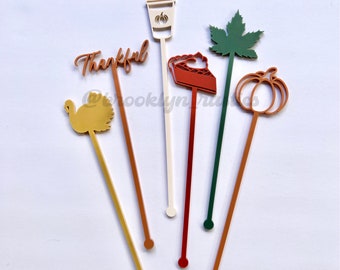 May include: Six colorful acrylic drink stirrers with various fall-themed designs, including a coffee cup, a pumpkin, a turkey, a maple leaf, a pie slice, and the word "Thankful".