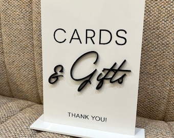 May include: A white acrylic sign with black lettering that reads "CARDS & Gifts" and "THANK YOU!". The sign is designed to stand upright on a white base, perfect for a wedding or event.