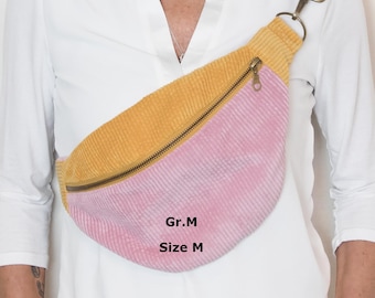May include: A corduroy crossbody bag featuring a mustard yellow upper section and a pink lower section, separated by a brass-toned zipper. The adjustable strap has a colorful geometric pattern. The bag is labeled with the text Gr.M and Size M.
