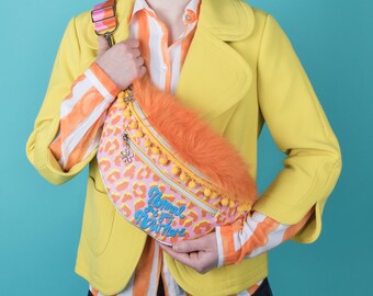 May include: A fanny pack featuring a pink and orange leopard print, a bright orange faux fur top panel, and yellow pom-pom trim. The bag displays the embroidered blue text "Normal is a setting on the dryer" and includes a colorful striped adjustable strap.