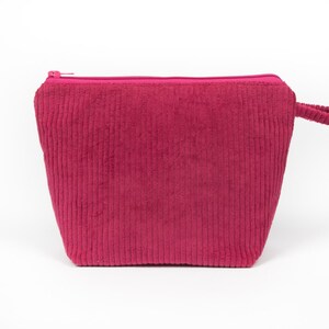 May include: A magenta corduroy cosmetic bag with a zip closure and a small handle. The bag has a rectangular shape with vertical ribbing. The background is white.