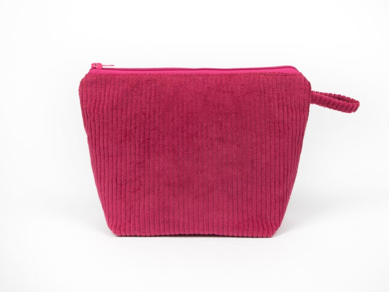 May include: A magenta corduroy cosmetic bag with a zip closure and a small handle. The bag has a rectangular shape with vertical ribbing. The background is white.