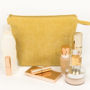May include: A mustard yellow corduroy cosmetic bag with a zip, alongside various beauty products. These include a lipstick, compact, foundation, and a jar of powder, all in shades of gold and beige.