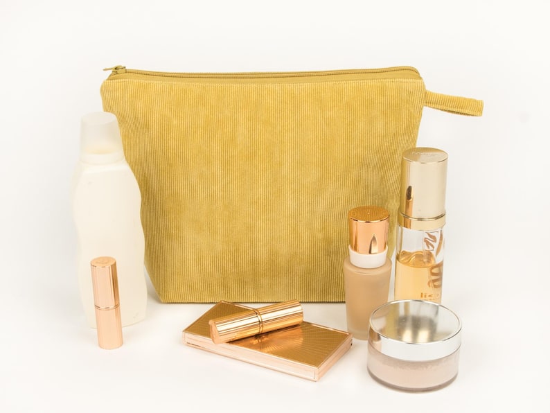 May include: A mustard yellow corduroy cosmetic bag with a zip, alongside various beauty products. These include a lipstick, compact, foundation, and a jar of powder, all in shades of gold and beige.