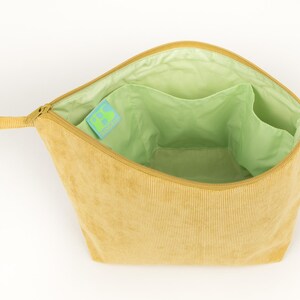 May include: Open, light yellow corduroy cosmetic bag. The interior is light green with multiple pockets. A small tag with a green and blue design is visible inside. The bag has a zip closure and a small loop handle. The bag's dimensions are not visible.