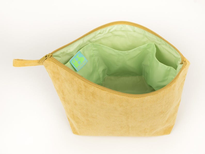 May include: Open, light yellow corduroy cosmetic bag. The interior is light green with multiple pockets. A small tag with a green and blue design is visible inside. The bag has a zip closure and a small loop handle. The bag's dimensions are not visible.