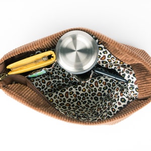 May include: A brown corduroy tote bag with a leopard print lining. The bag is open and contains a yellow wallet, a silver water bottle, and a black phone.